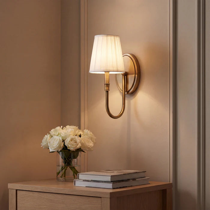 A wall-mounted sconce with a white pleated drum shade and a curved brass-toned arm, shown lit on a beige wall above a wooden console table with white roses and stacked books.